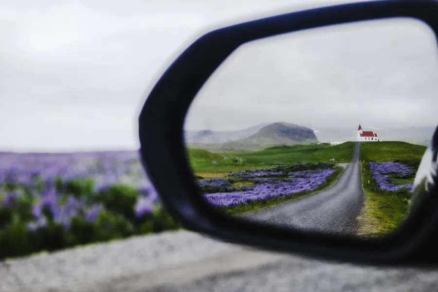 A red-roofed church, road and lupine fields in Iceland, framed in a car's wing mirror.