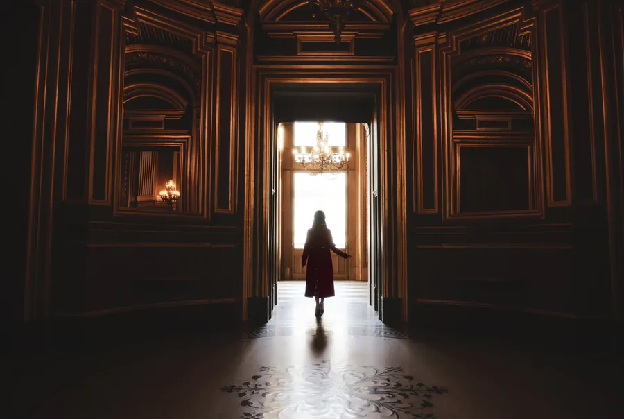 A silhouette in the doorway of a dark, panelled Paris hall lit by a chandelier.